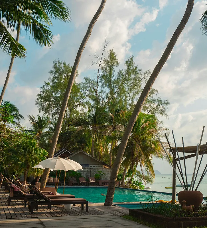 Resort pool area with lounge chairs, an umbrella, and palm trees under a partly cloudy sky near the ocean.