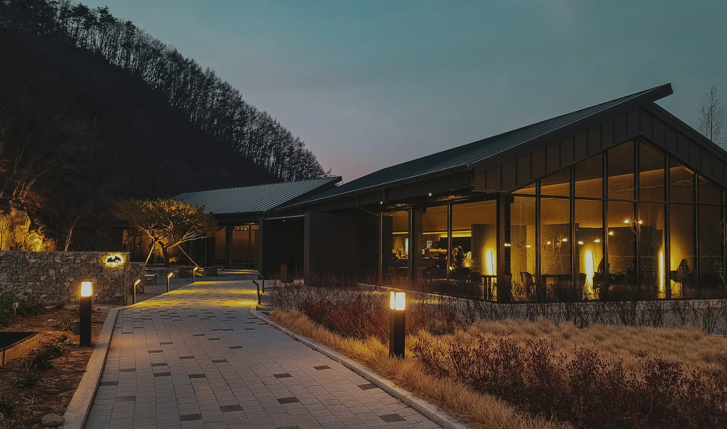Modern glass building illuminated by warm lights along a paved pathway at dusk with trees and hills in the background.