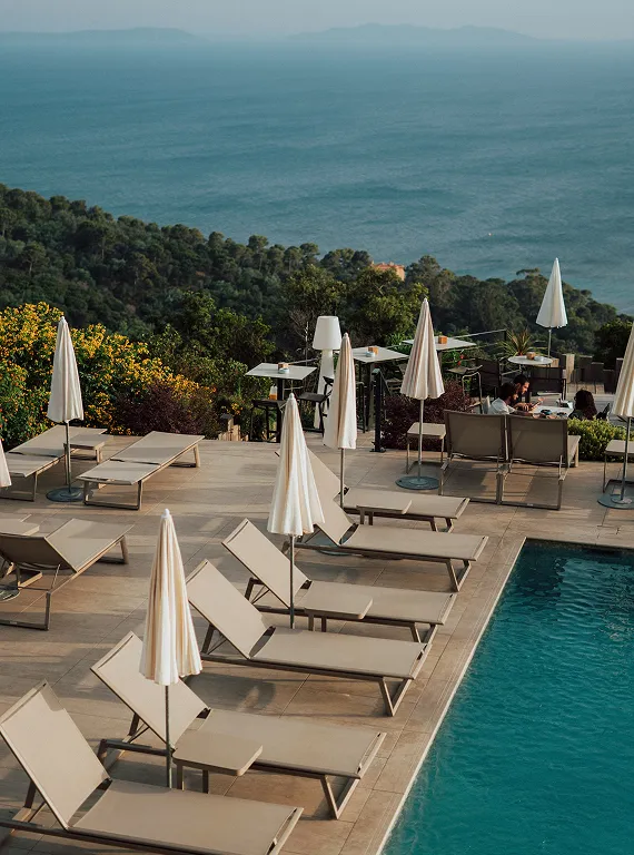 Poolside area with beige lounge chairs and closed umbrellas overlooking a forested hillside and ocean.