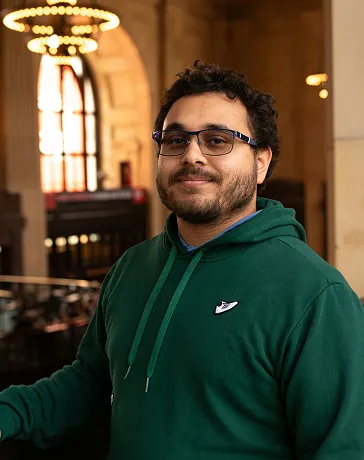 Man with glasses and curly hair wearing a green hoodie, standing indoors with arched windows and chandelier lights in the background.