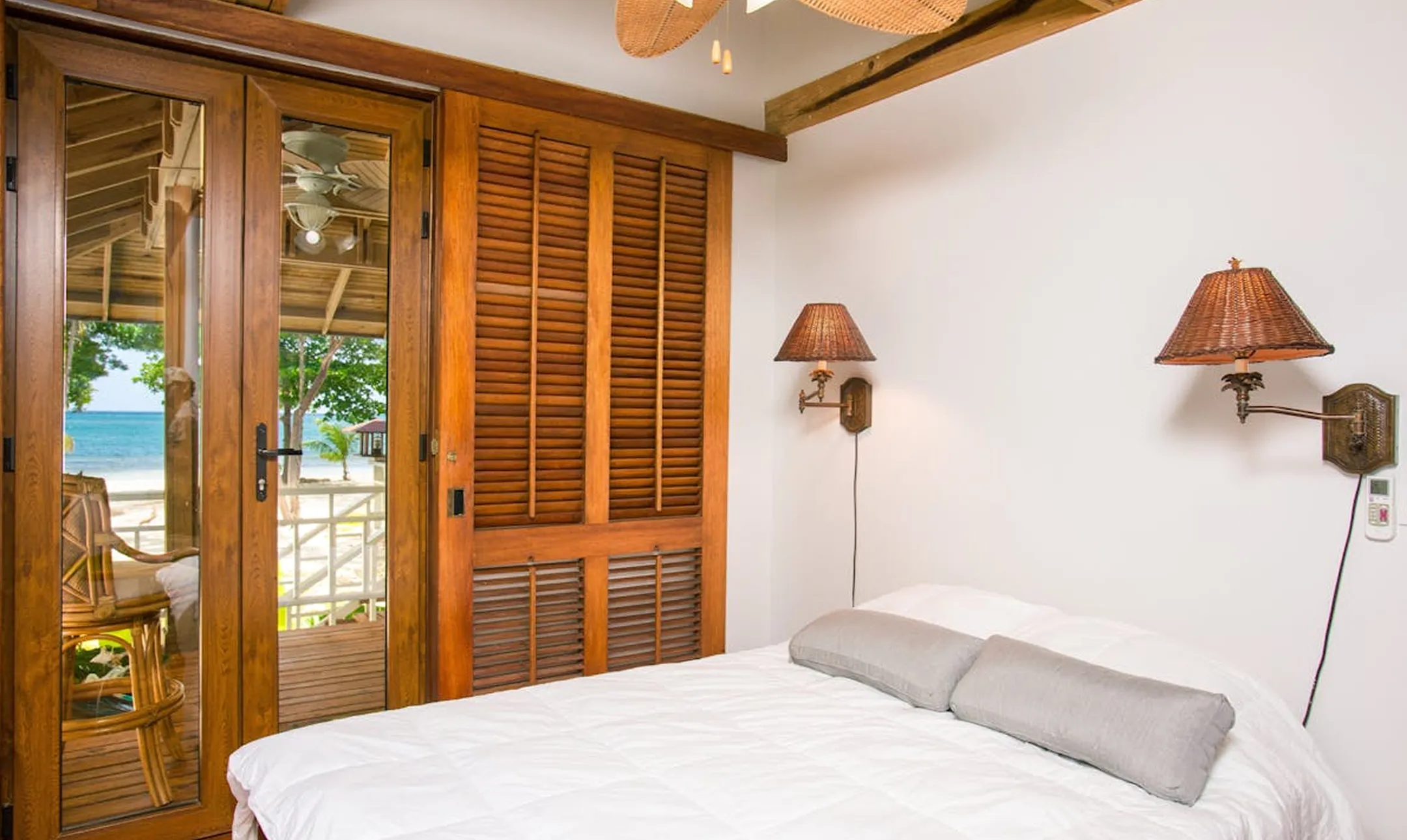 Bedroom with white bedding, two gray pillows, wooden sliding closet doors, wall-mounted wicker lamps, and glass doors opening to a balcony overlooking a beach and ocean.