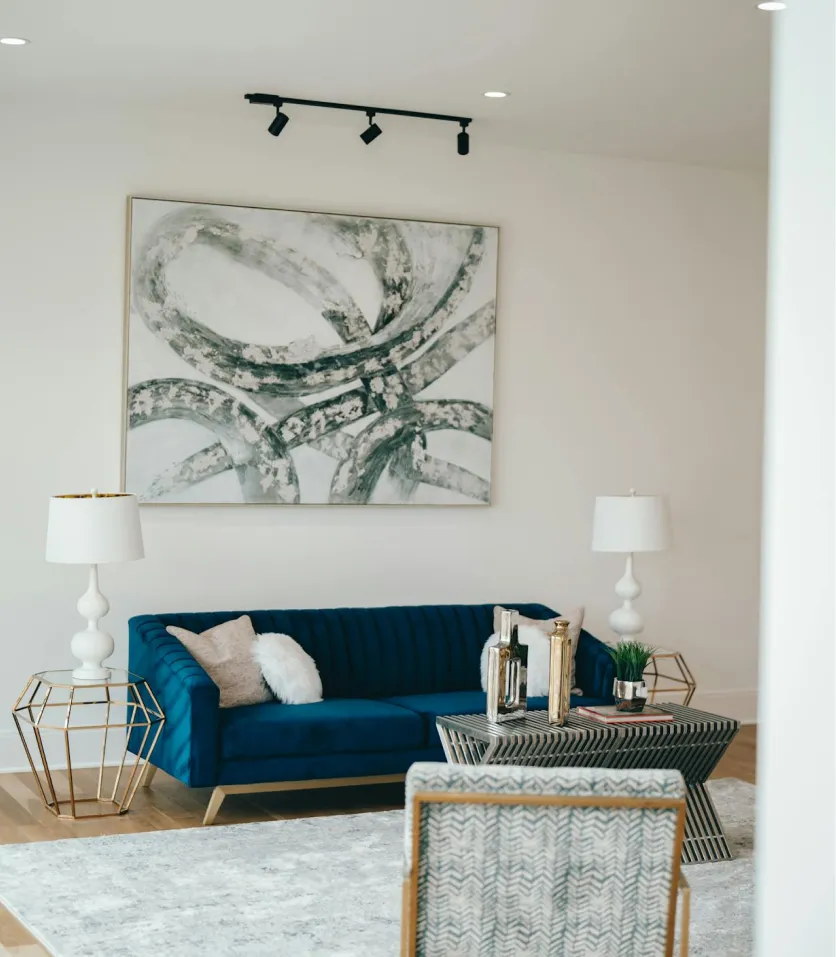 Modern living room with a blue velvet sofa, abstract wall art, geometric side tables with white lamps, and a patterned chair.
