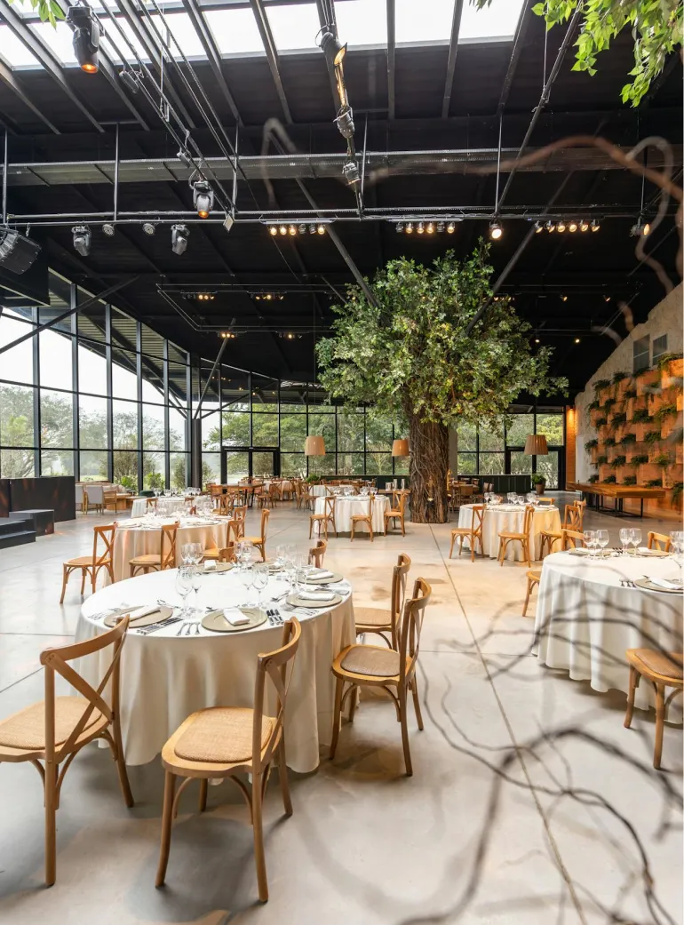 Spacious modern dining area with round tables covered in white tablecloths and wooden chairs surrounding a large artificial tree in the center.