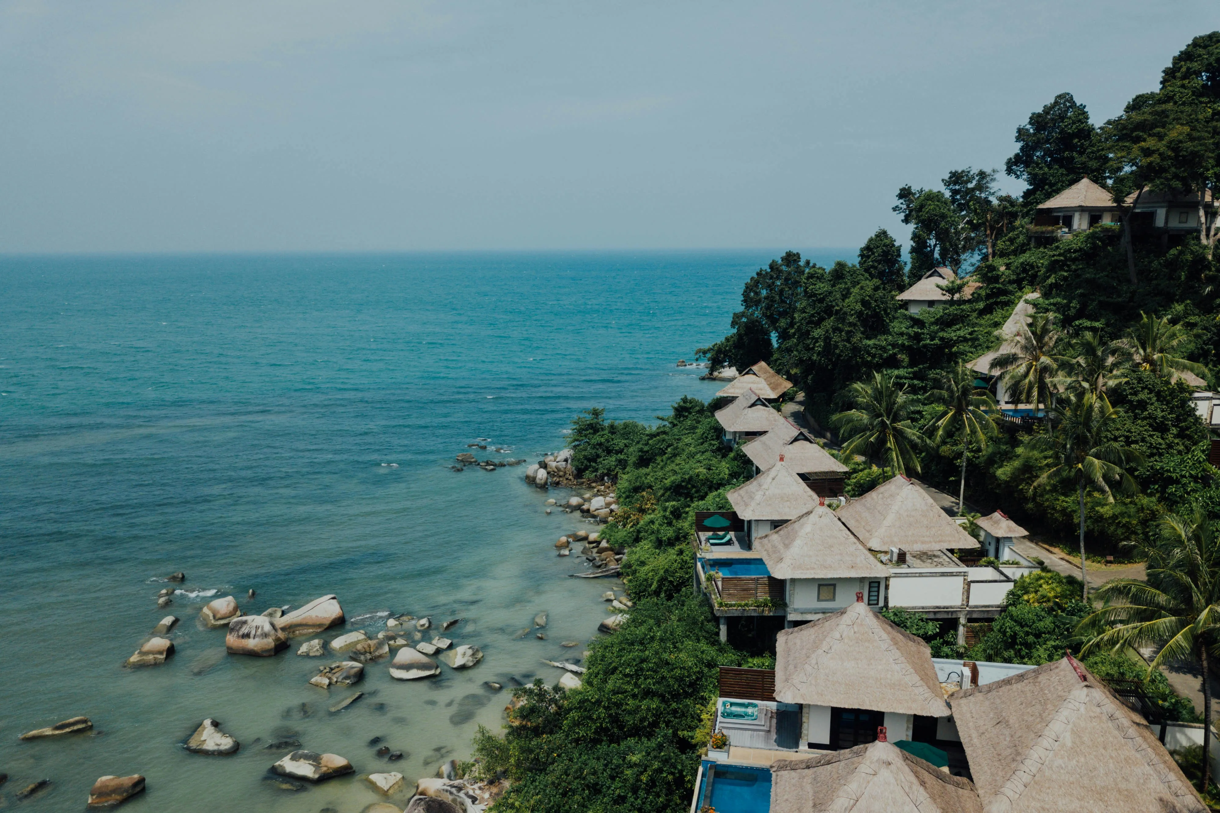Coastal resort with thatched-roof villas surrounded by lush greenery overlooking blue ocean waters and rocky shoreline.