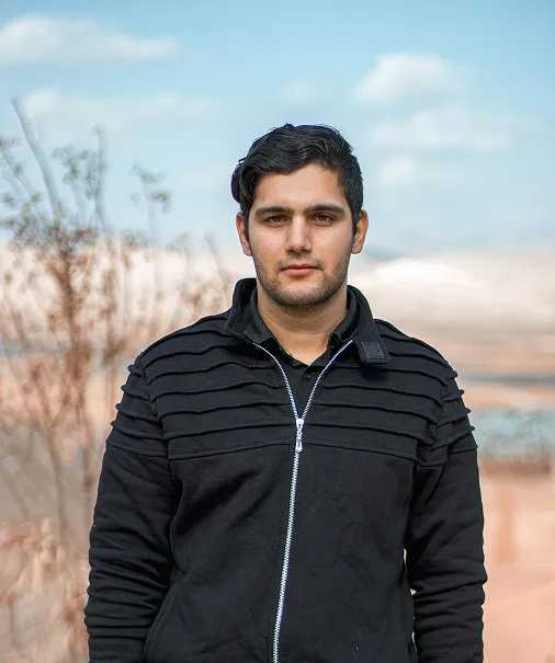 Young man with dark hair wearing a black zip-up sweater standing outdoors with blurred trees and a cloudy sky in the background.