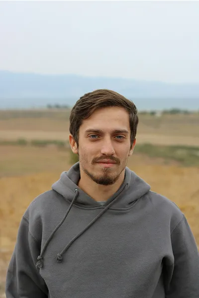 Young man with short brown hair and beard wearing a gray hoodie standing outdoors with a blurred landscape background.