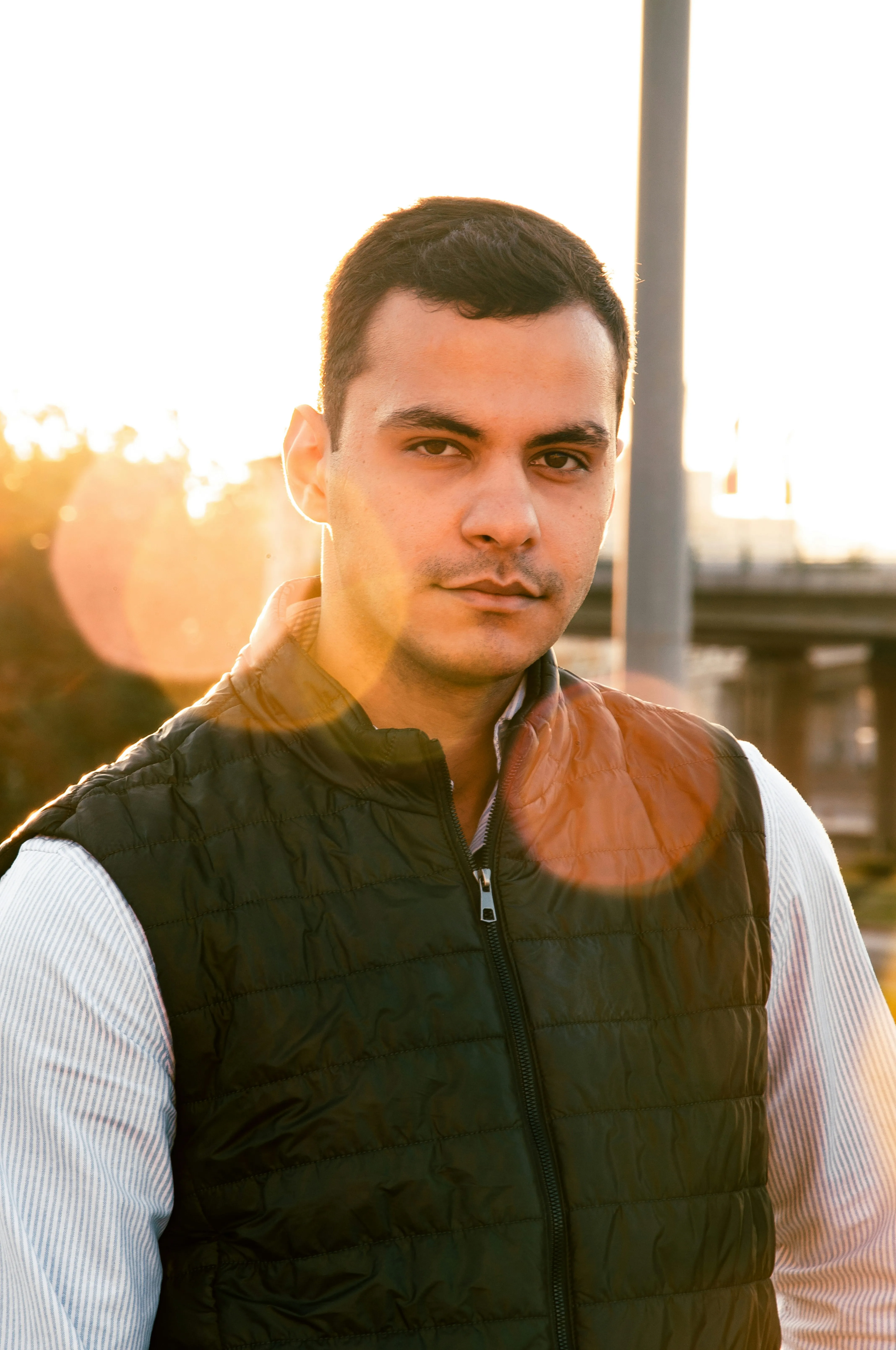 Portrait of a man wearing a black quilted vest and light striped shirt standing outdoors at sunset with lens flare.