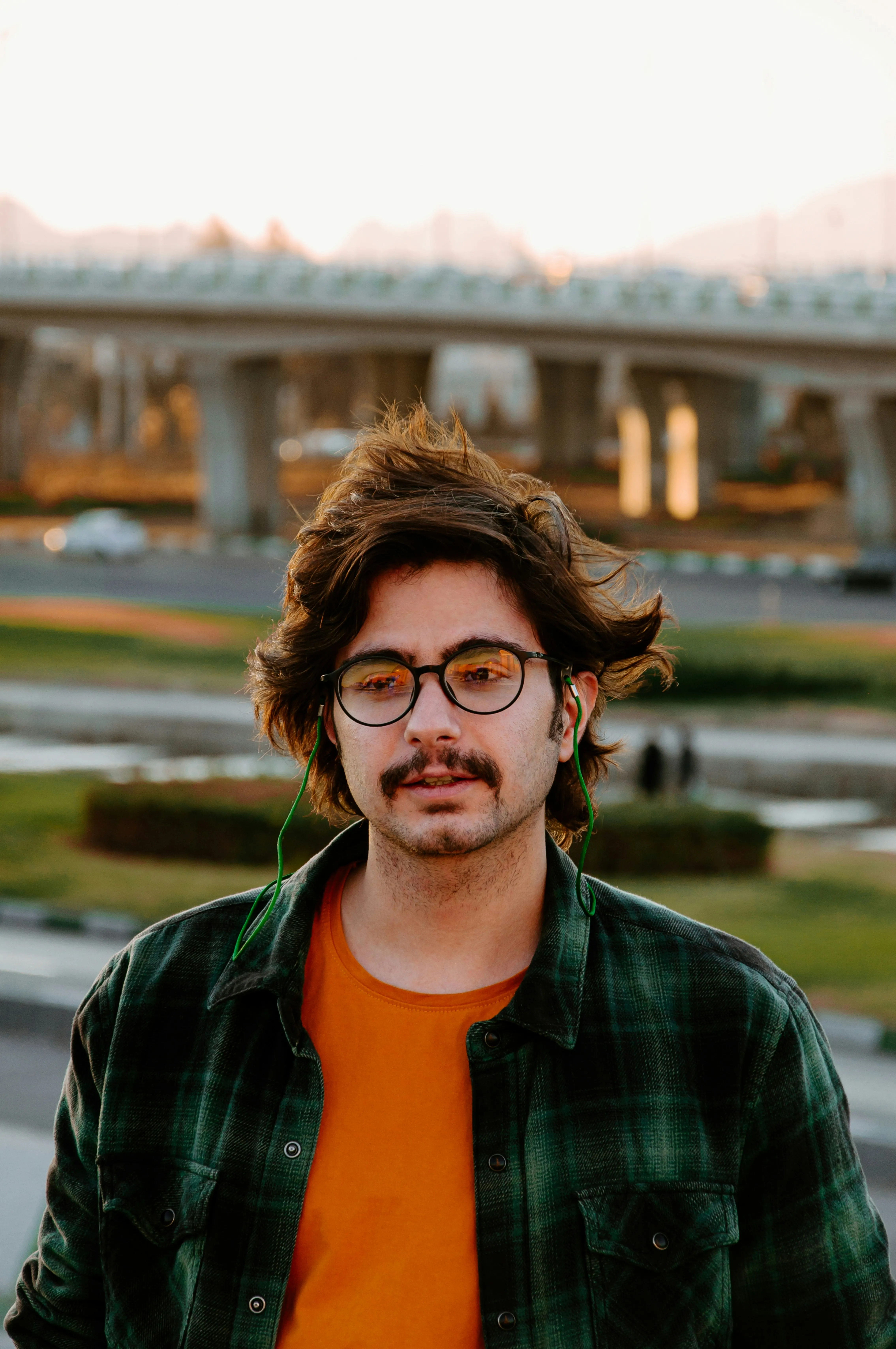 Young man with glasses and mustache wearing a green plaid jacket and orange shirt outdoors near a road and bridge at sunset.