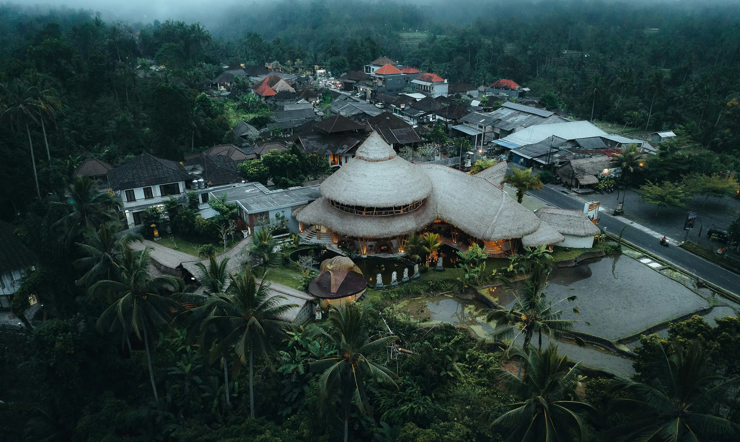 Aerial view of a tropical village with a large thatched round building surrounded by palm trees and rice fields in a misty landscape.