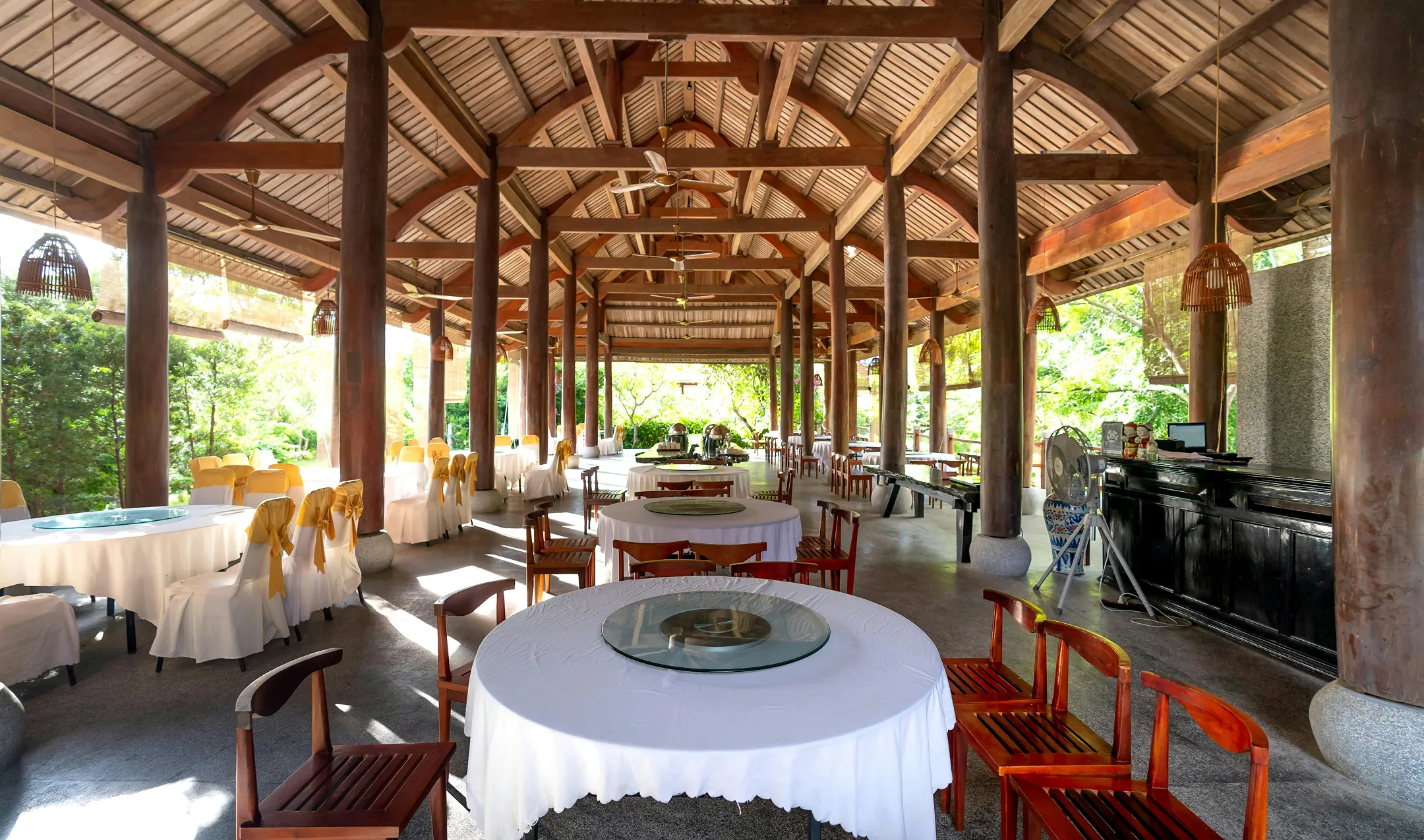 Open-air restaurant with round tables covered in white cloth and wooden chairs under a wooden beam roof with hanging lanterns.