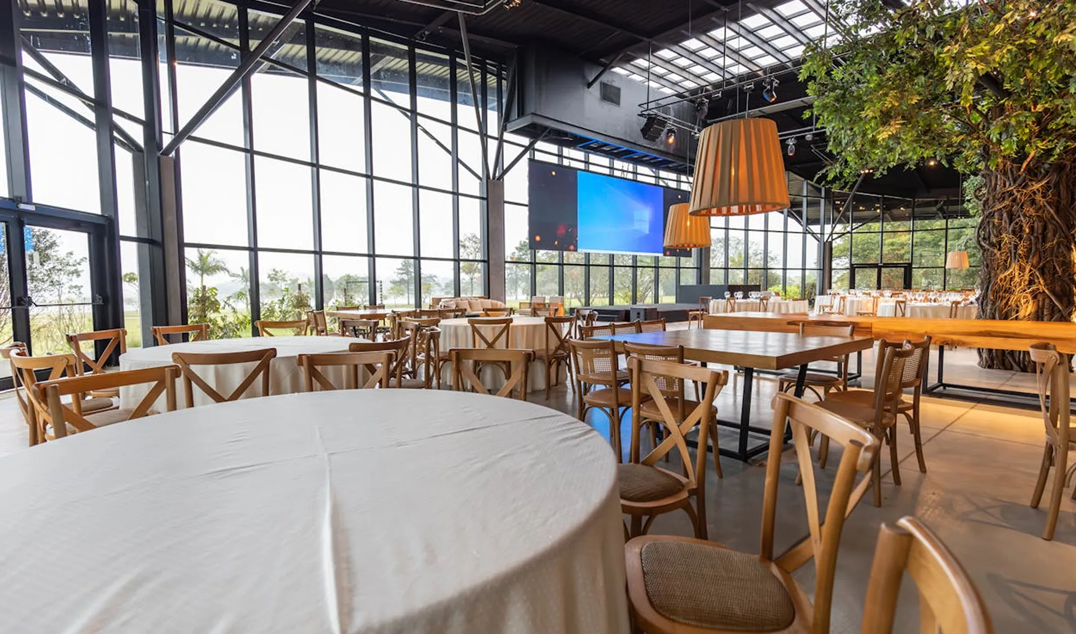 Spacious modern event hall with round and rectangular tables covered in white tablecloths and wooden chairs, large windows, pendant lights, and a large screen.