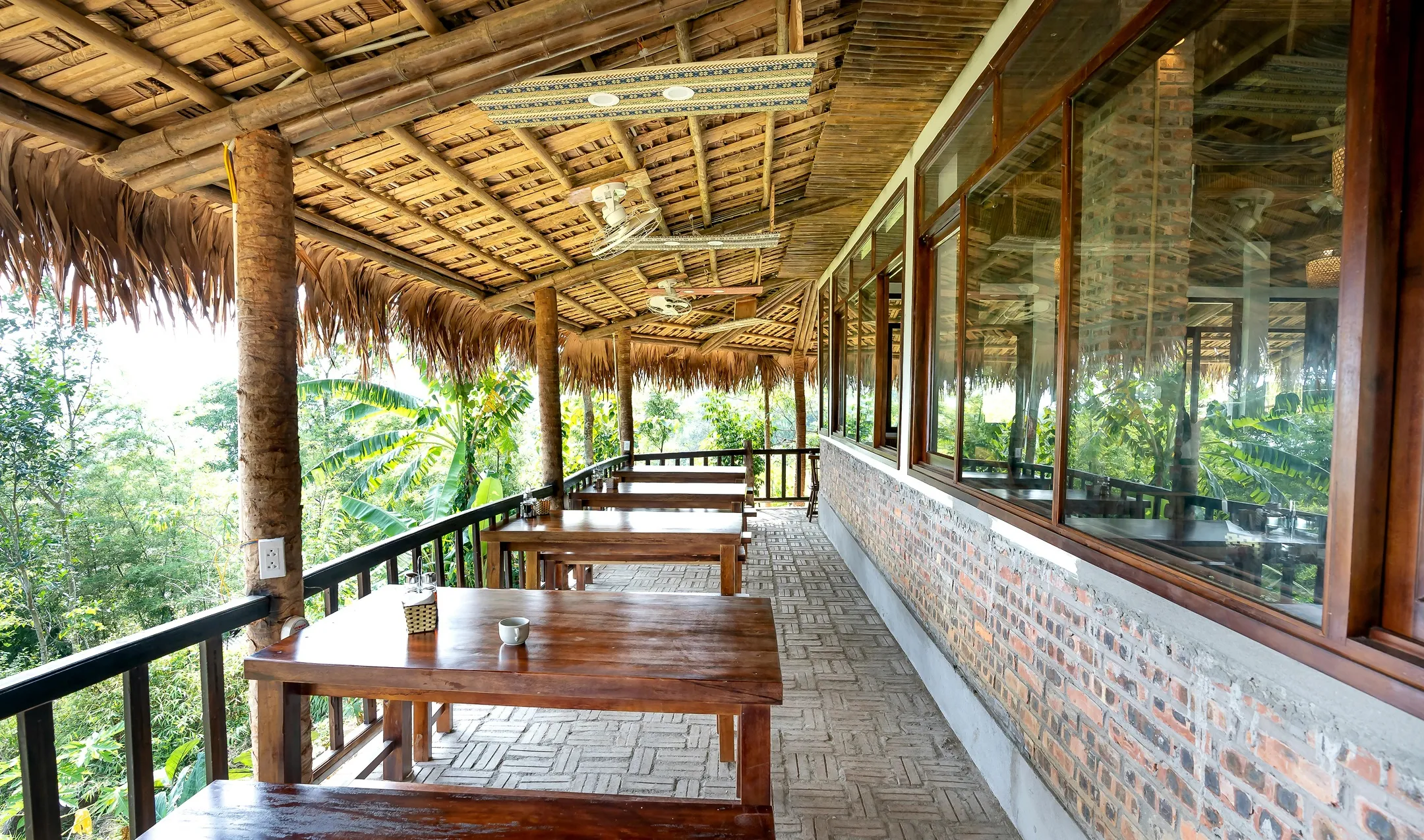 Outdoor covered wooden dining area with picnic tables overlooking green tropical foliage, featuring a thatched roof and brick wall with large windows.