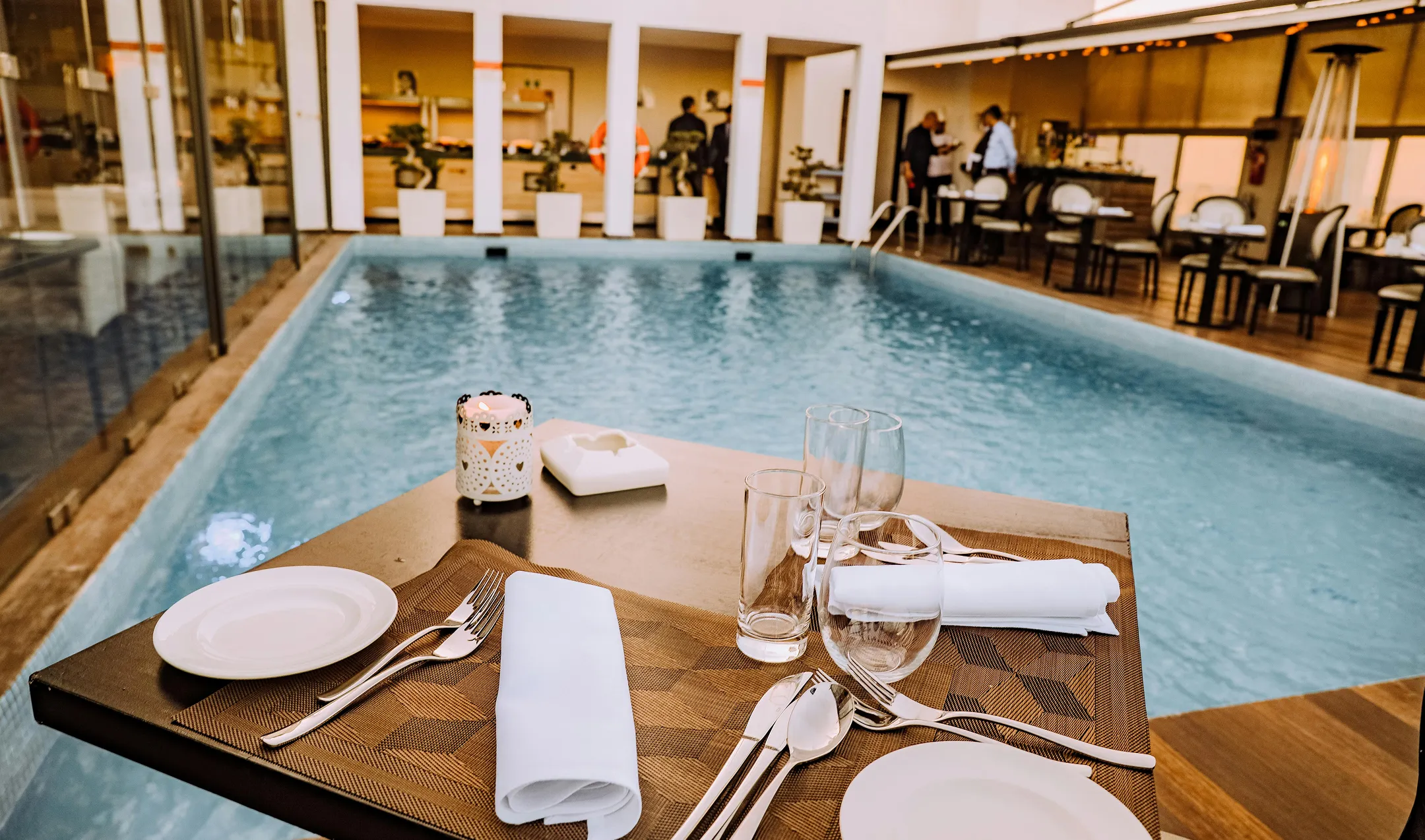 Table set with plates, cutlery, glasses, and napkins beside an indoor pool in a restaurant.
