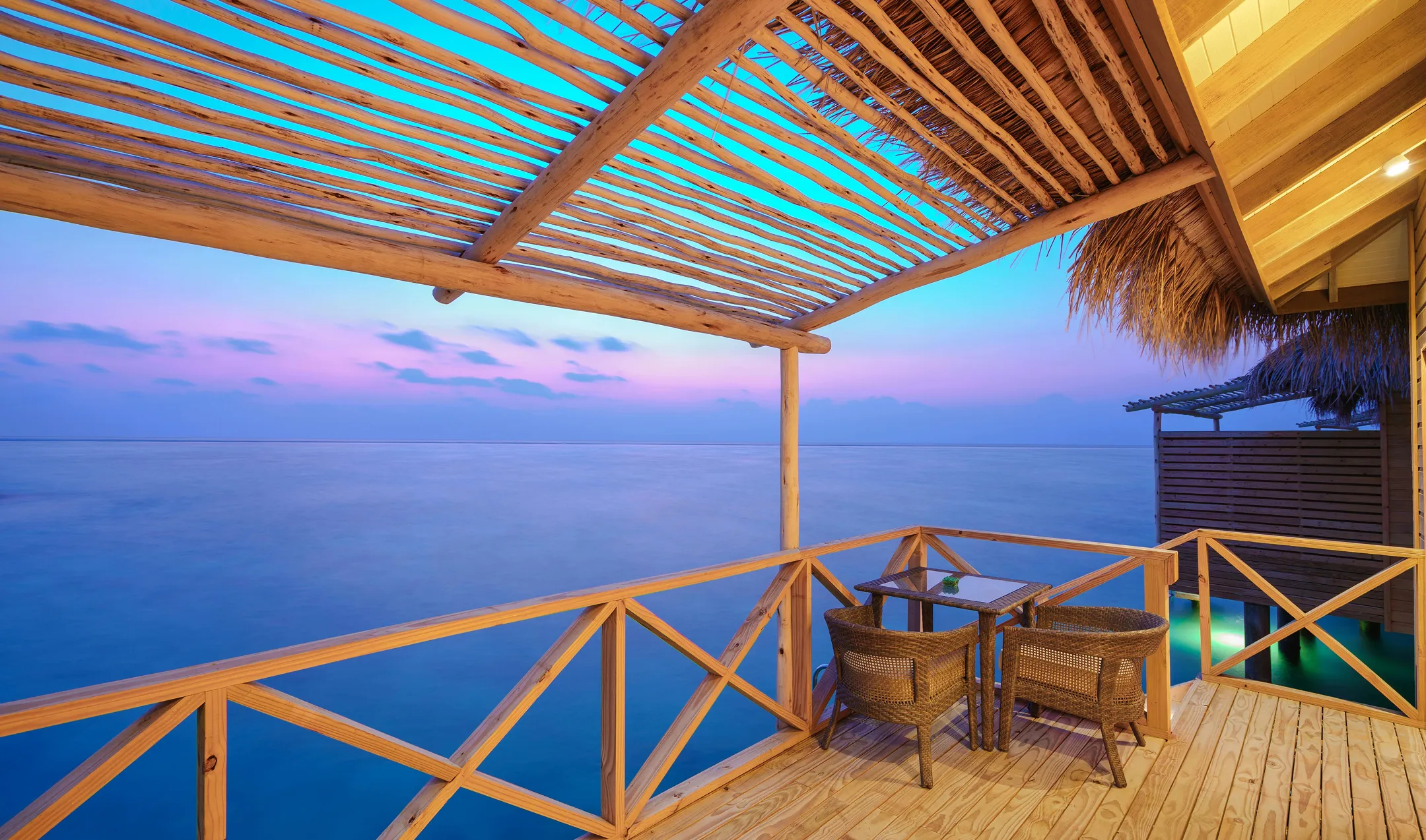 Wooden deck with two wicker chairs and a table overlooking calm ocean water at sunset under a wooden pergola.