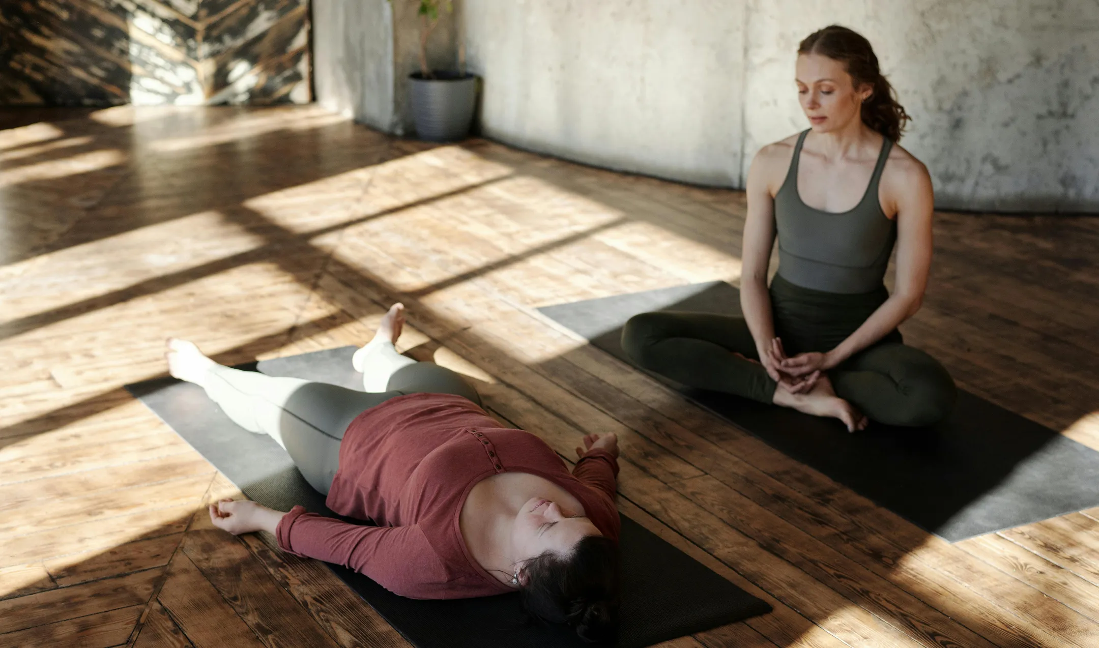 Two women practicing yoga in a sunlit room, one lying on a mat with eyes closed, the other sitting cross-legged nearby.