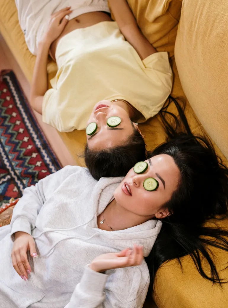 Two women lying on a yellow couch with cucumber slices on their eyes, relaxing and enjoying a spa treatment at home.