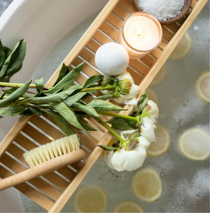 Bath tray with a lit candle, bath bomb, white flowers, and a wooden brush over a bathtub with lemon slices in the water.