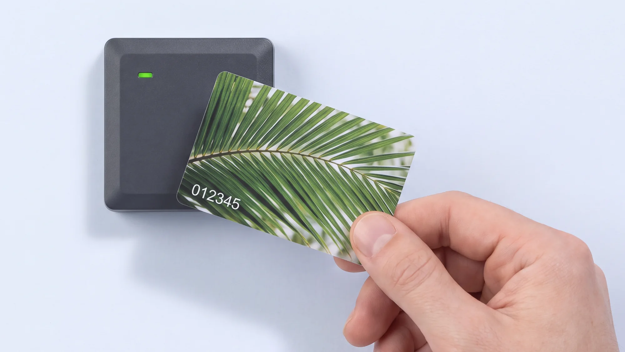 A hand presenting an RFID card to a matte dark grey wall reader. The card is printed edge to edge with a green palm frond and carries a serial number.