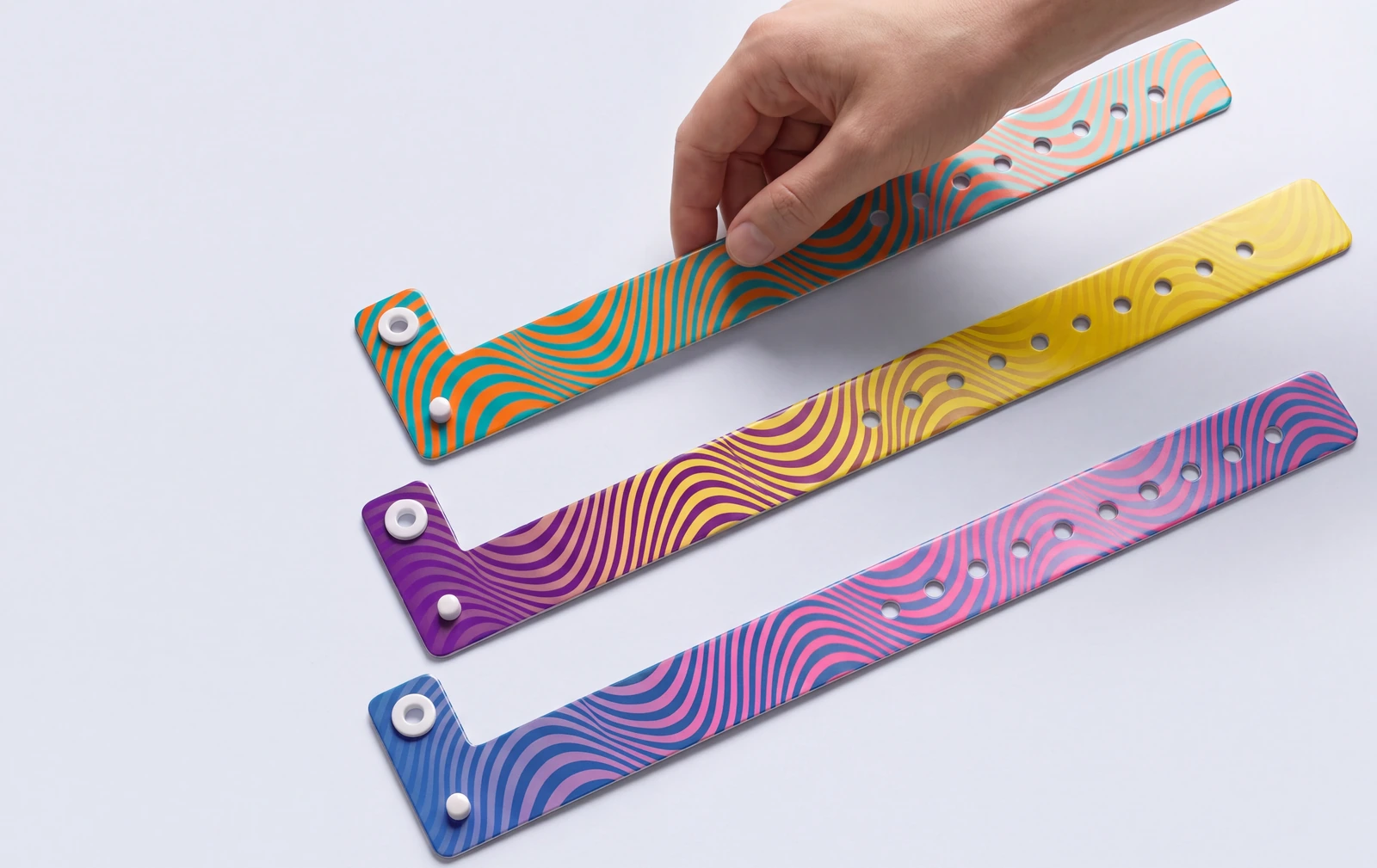 Three printed vinyl event wristbands with white snap studs and a row of adjustment holes, one being lifted by hand