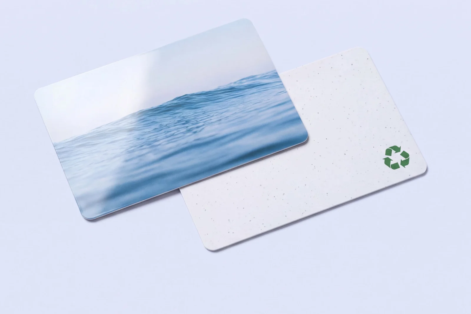 A gloss PVC card printed with an ocean photograph beside a speckled recycled PVC card marked with a green recycling symbol