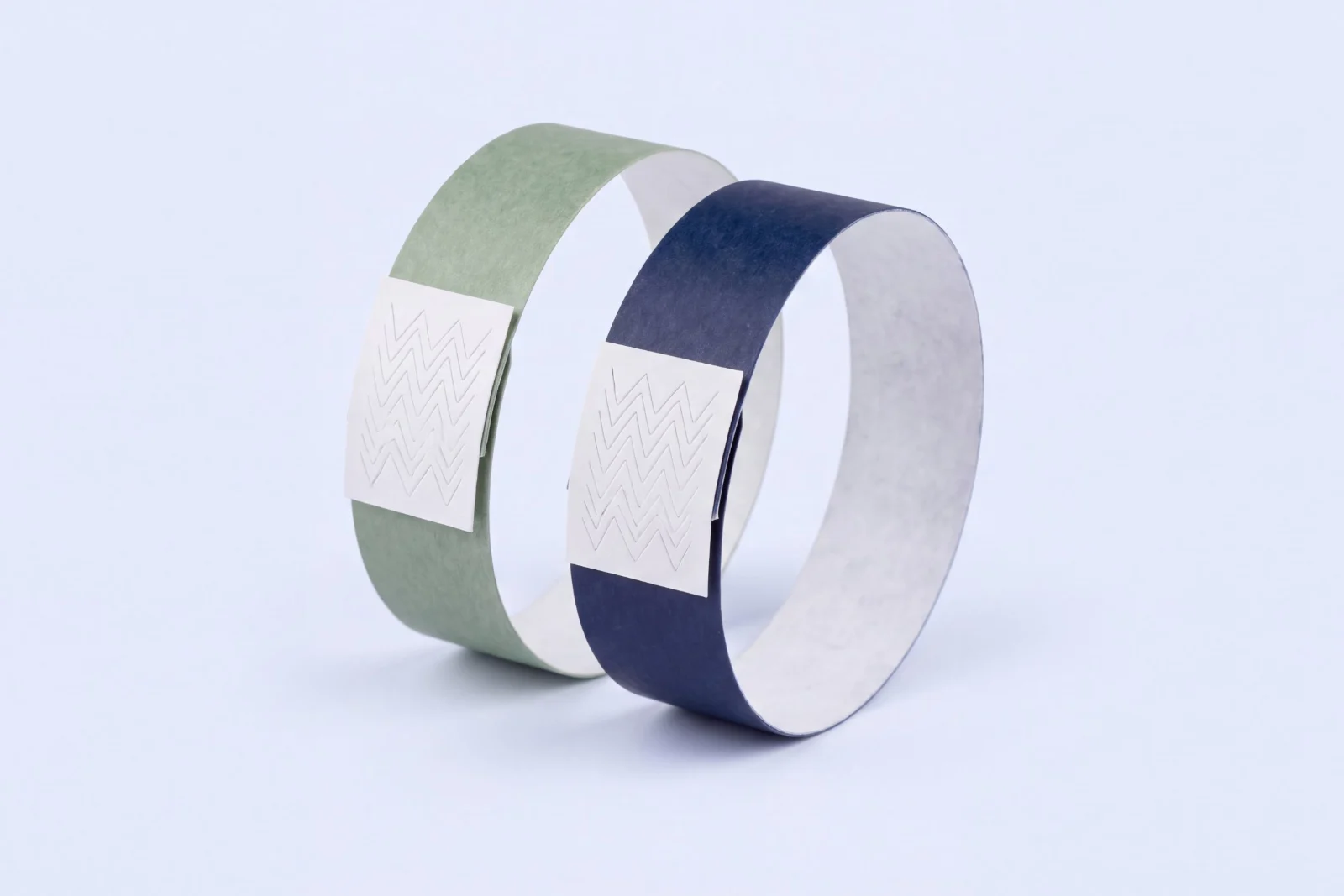 Two Tyvek wristbands in sage green and navy, each closed with a white tamper evident tab with an embossed zigzag pattern