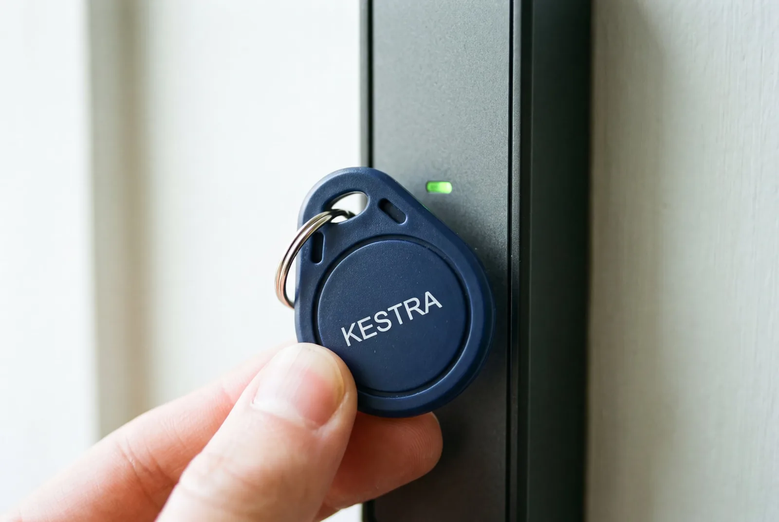 Fingertips holding a navy blue RFID key fob printed KESTRA on a split ring against a black wall reader with a green light