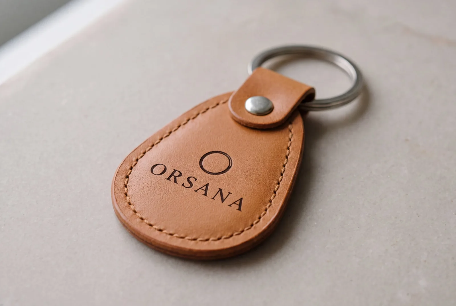 A tan leather RFID key fob with edge stitching and a riveted tab on a split ring, printed ORSANA, lying on pale stone