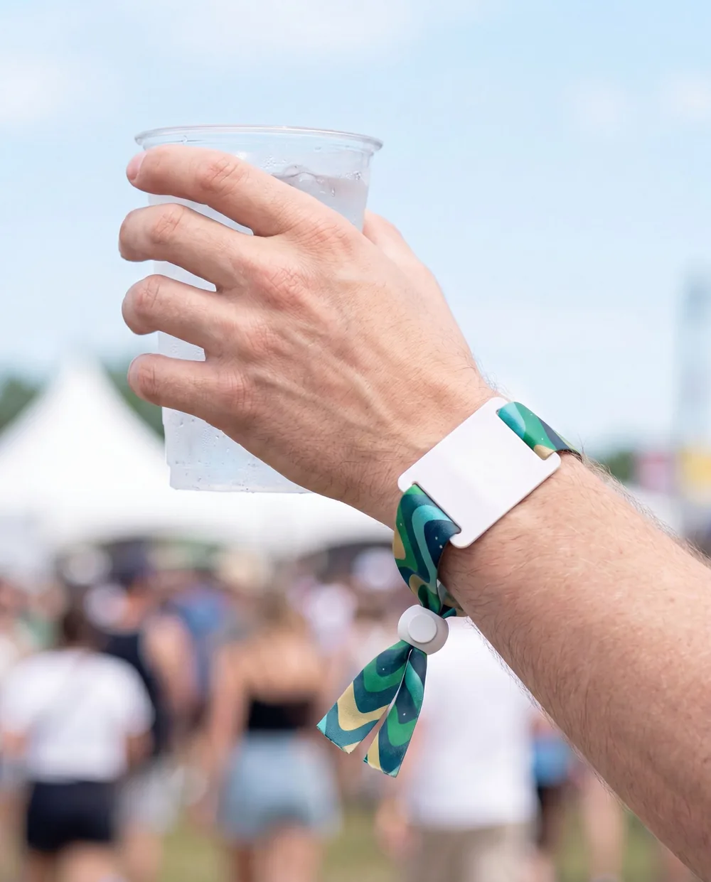 Raised hand holding a cup of water at a summer festival, wearing a printed fabric RFID wristband with a white tag