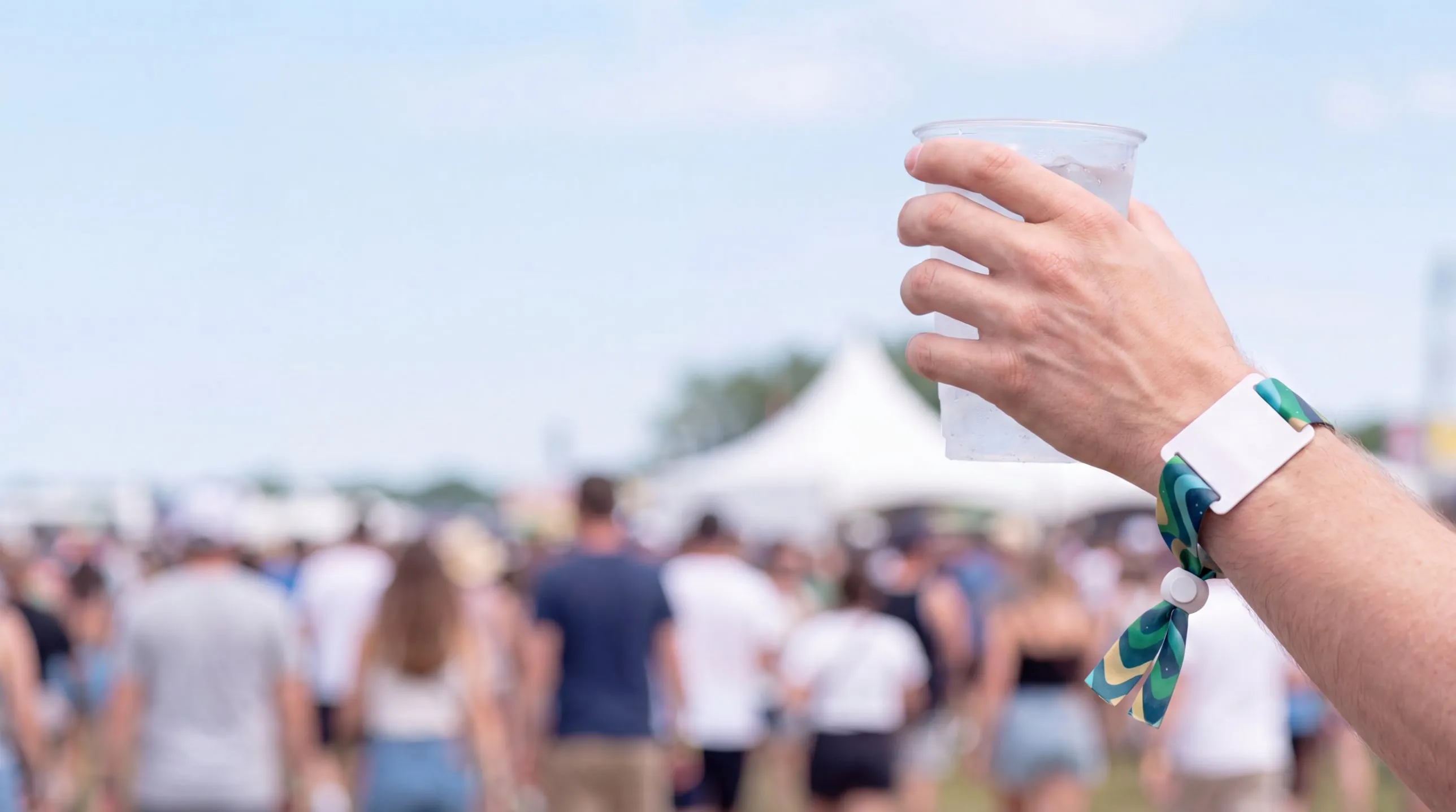 Raised hand holding a cup of water at a summer festival, wearing a printed fabric RFID wristband with a white tag, crowd and tents blurred behind