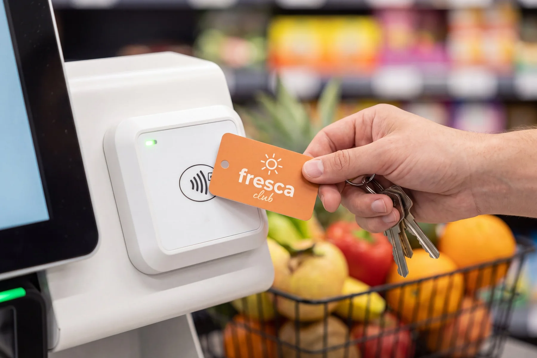 A hand tapping an orange loyalty key tag for a fictional grocery club on the contactless reader of a supermarket self-checkout, with a basket of fruit behind
