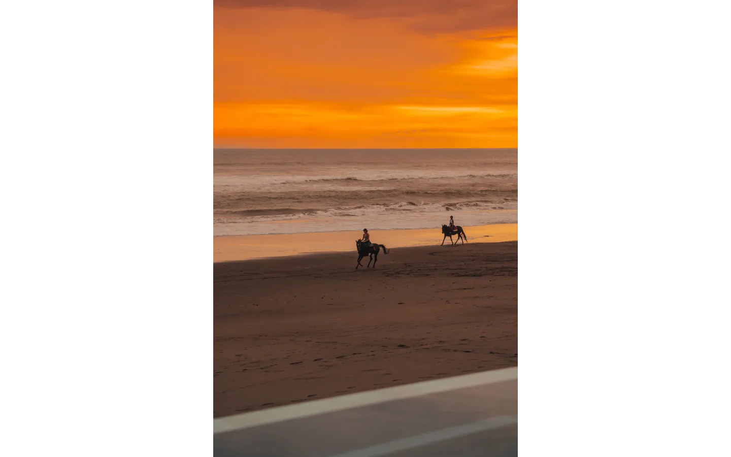 Two people riding horses along the beach at sunset with an orange sky over the ocean.