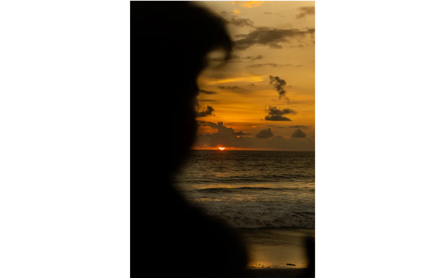 Silhouette of a person watching a sunset over the ocean with an orange sky and scattered clouds.