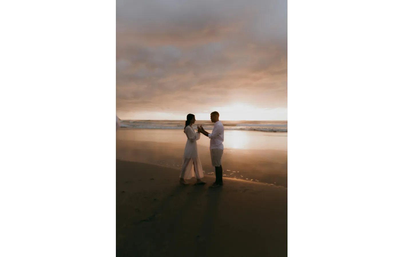 Couple standing on a beach holding hands facing each other at sunset with cloudy sky.