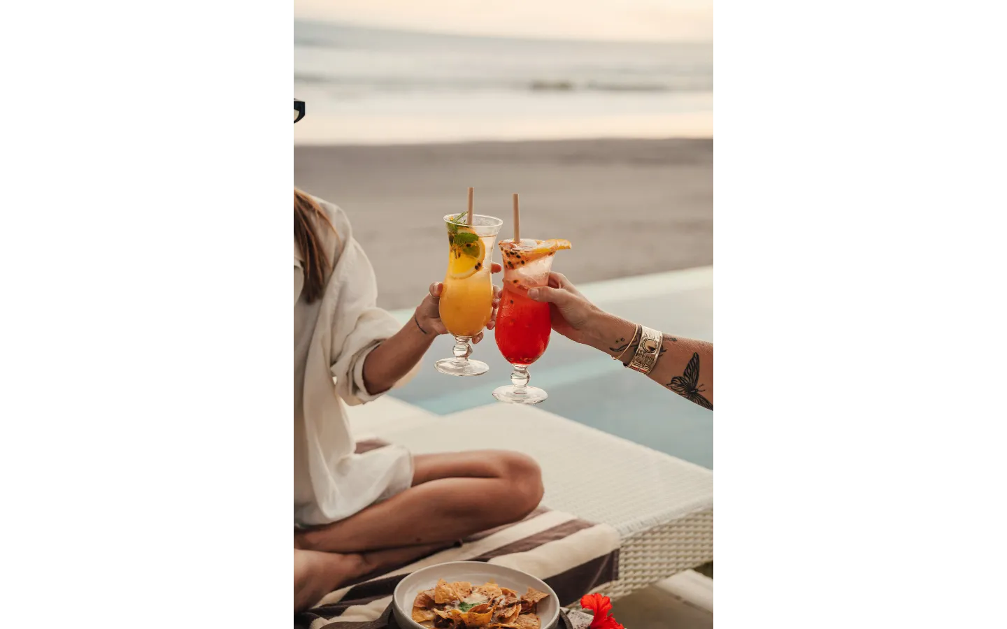 Two people clinking tropical cocktails by the beach during sunset, with a bowl of chips nearby.