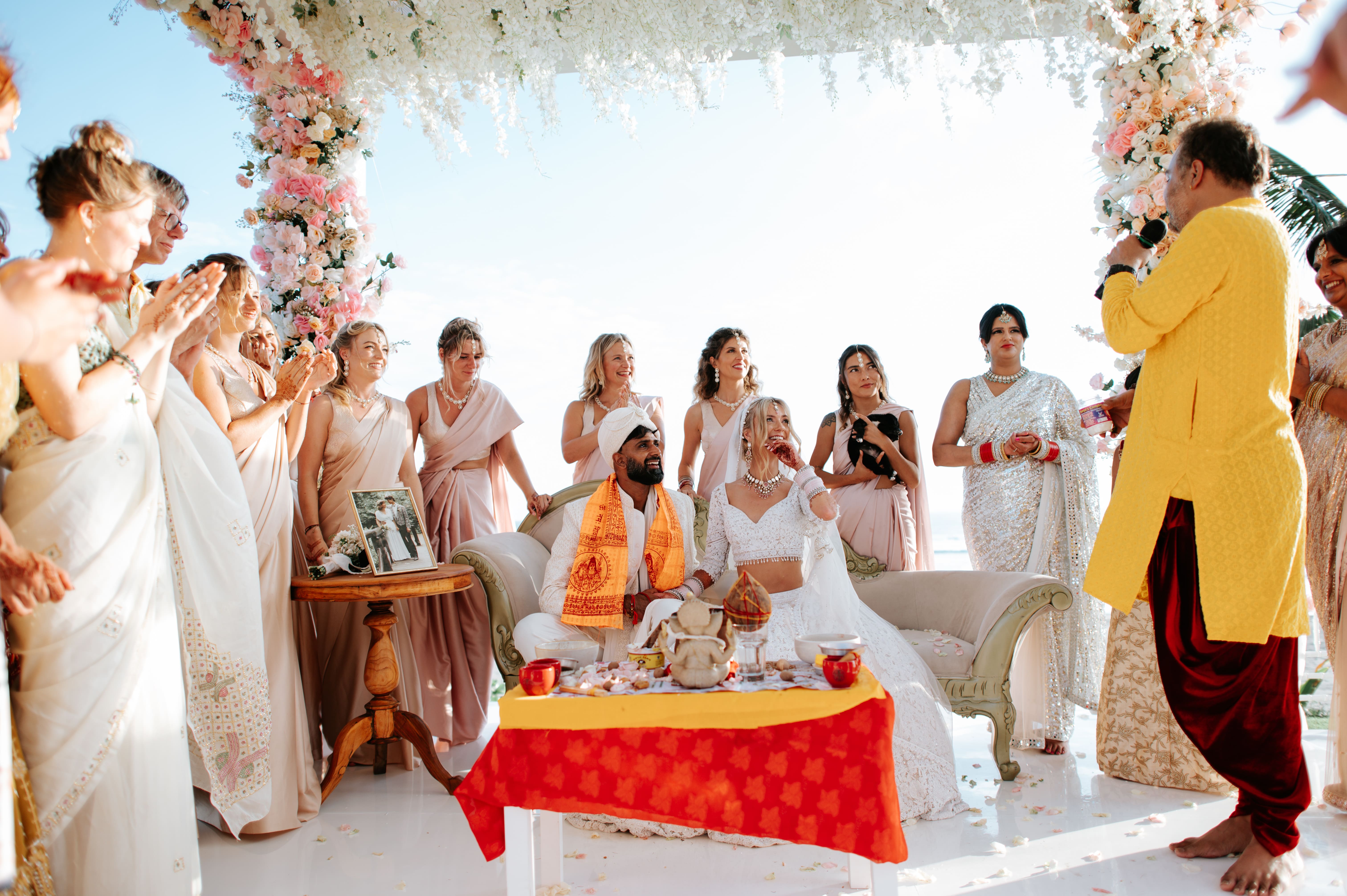 Bride and groom seated under a floral arch during a wedding ceremony surrounded by smiling guests.