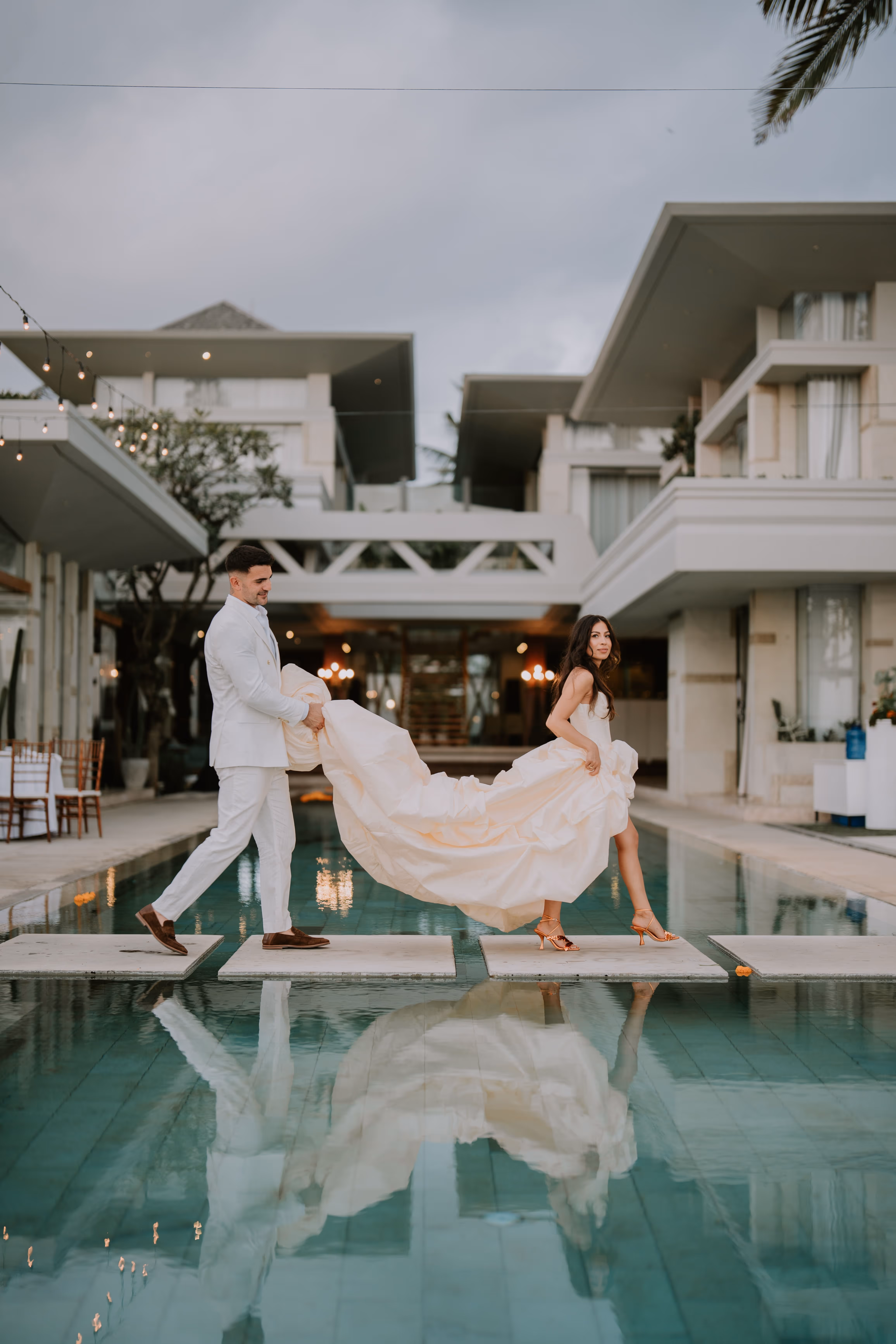 Bride in a flowing gown and groom in a white suit walking on stone slabs over a pool at modern villa.