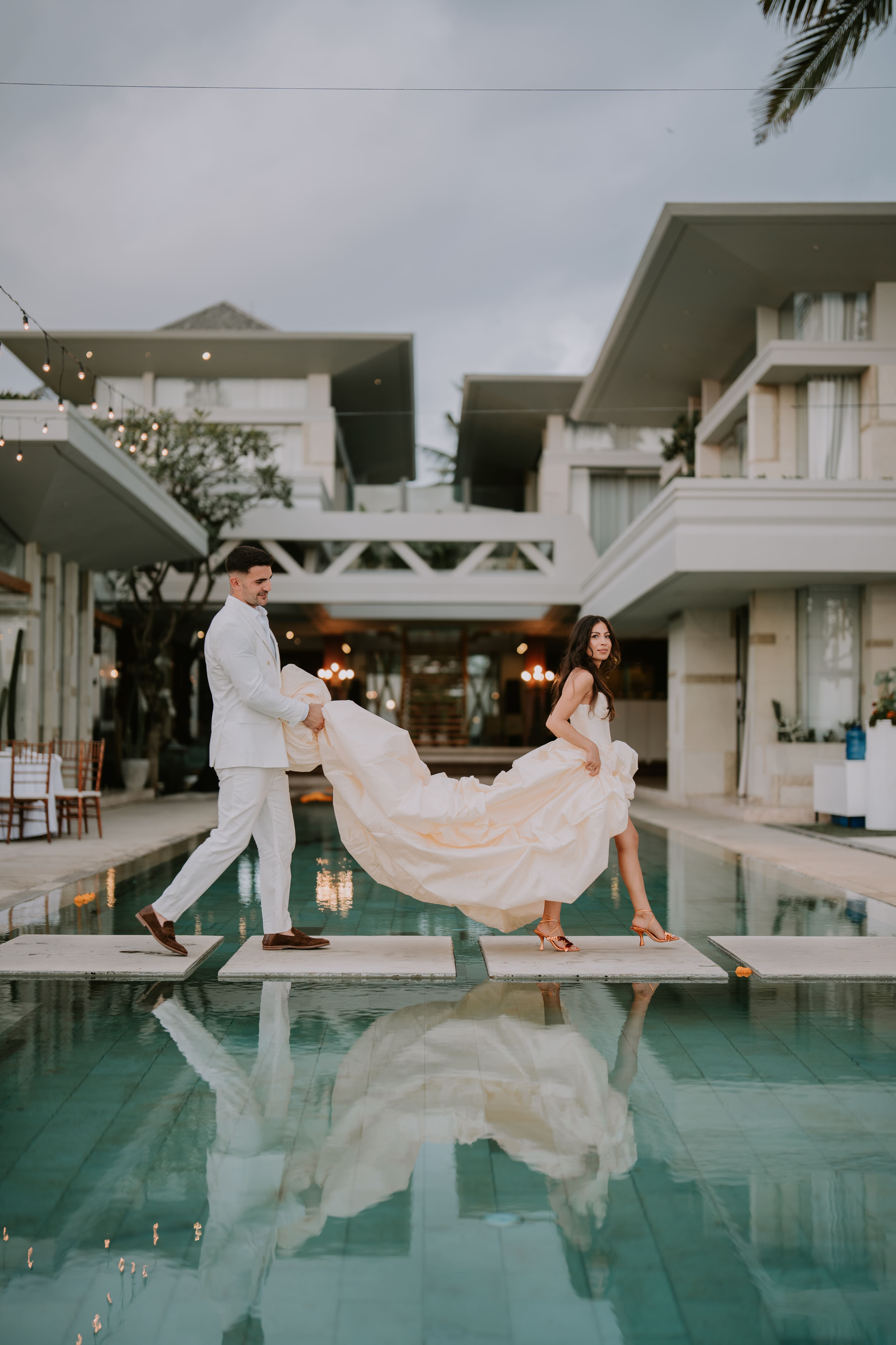 Bride in a flowing gown and groom in a white suit walking on stone slabs over a pool at modern villa.