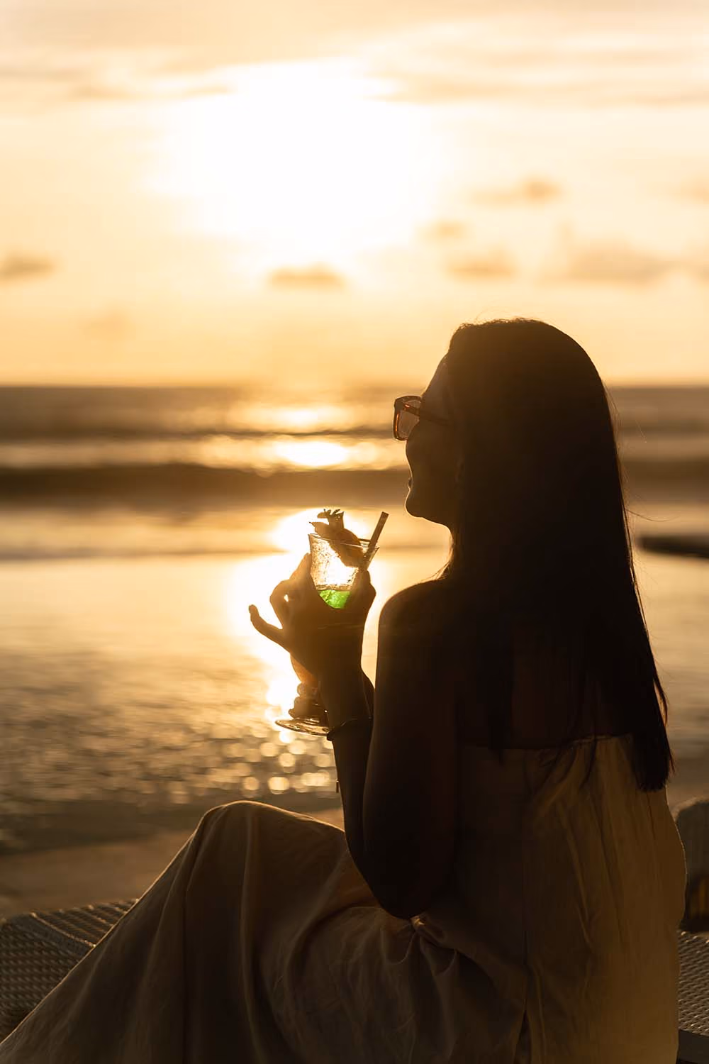 Woman wearing glasses sitting by the beach at sunset, holding a green cocktail with a straw and garnish.