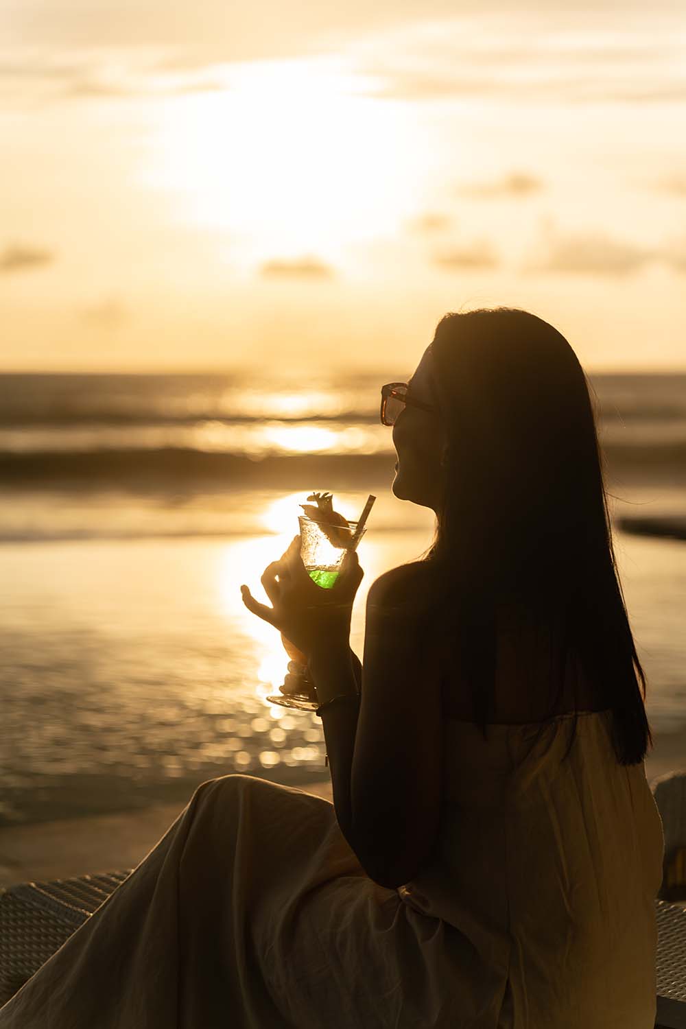 Woman wearing glasses sitting by the beach at sunset, holding a green cocktail with a straw and garnish.