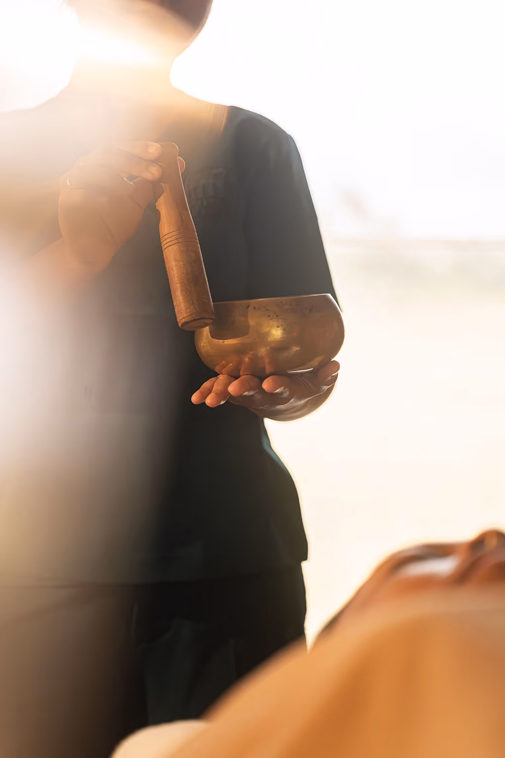 Person holding a singing bowl and wooden mallet with warm sunlight in the background.