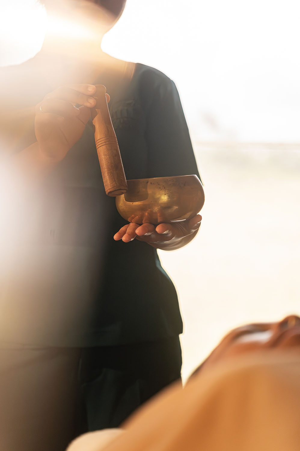 Person holding a singing bowl and wooden mallet with warm sunlight in the background.