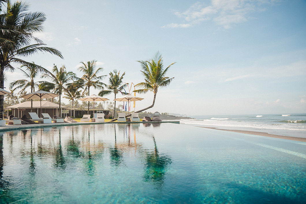 Infinity pool reflecting palm trees and umbrellas with sandy beach and ocean in background under clear sky.