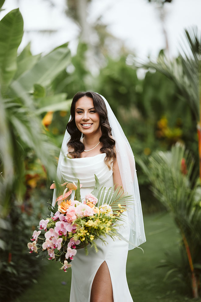 Smiling bride in white dress holding a colorful bouquet in a lush green outdoor setting.