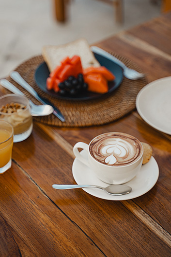 Cup of hot chocolate with latte art on a white saucer and spoon, next to a plate of toast and fruit on a wooden table.