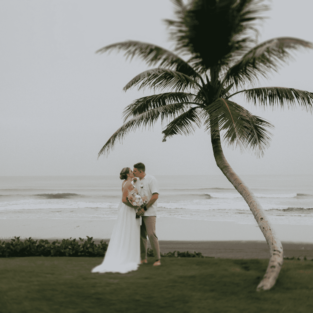 Bride and groom kissing on a beach with a palm tree and ocean waves in the background.