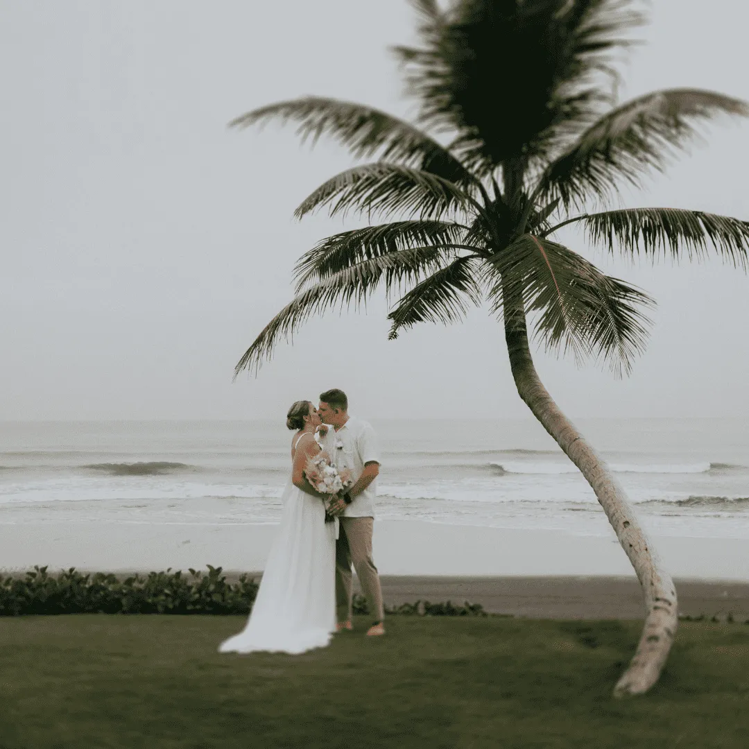 Bride and groom kissing on a beach with a palm tree and ocean waves in the background.