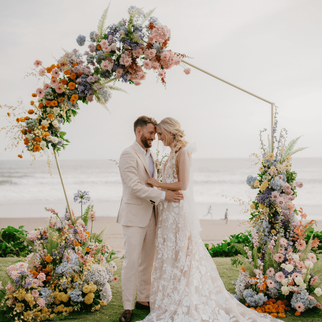 Bride and groom embracing under a floral arch on a beach during wedding ceremony.