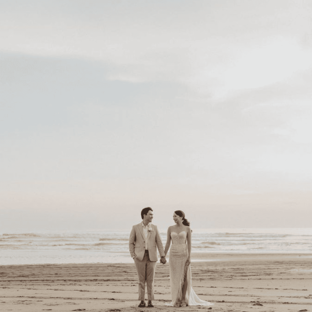 Bride and groom holding hands and smiling on a beach with ocean waves in the background.