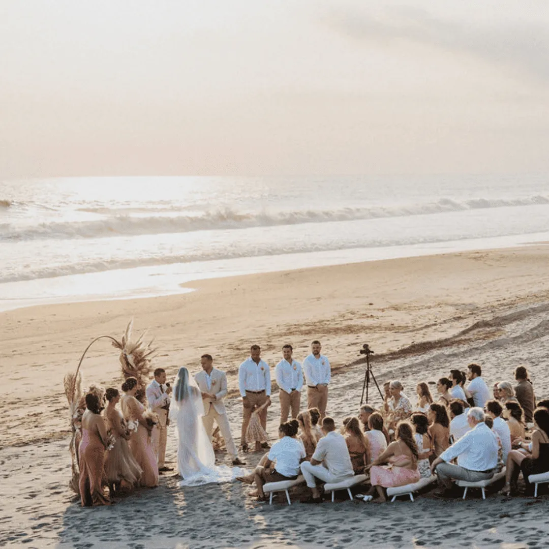Couple exchanging vows during a beach wedding ceremony at sunset with guests seated nearby.