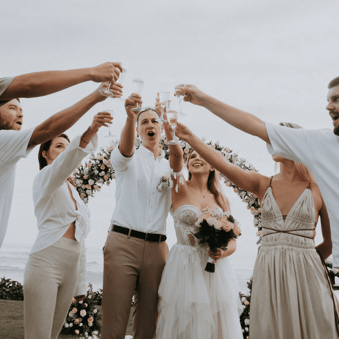 Bride and groom raising glasses for a toast with guests outdoors at a floral wedding arch.